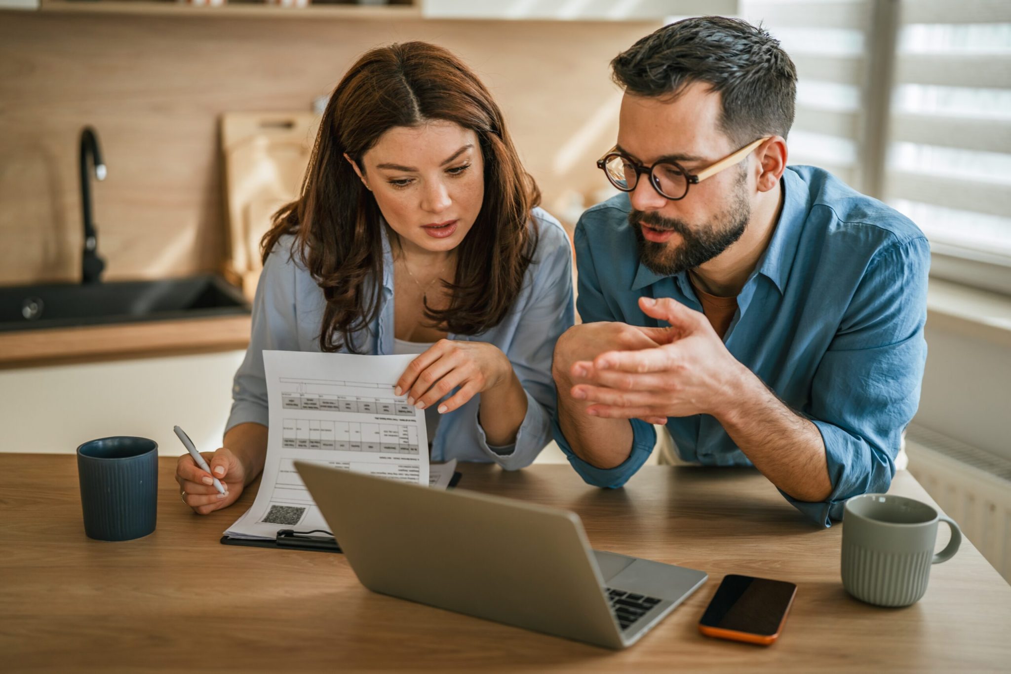 Young couple managing household finances, reviewing documents, and using a laptop to plan expenses and investments while working together in their modern kitche