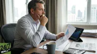 A man in a light blue shirt sits at a wooden desk, holding a printed graph showing a downward trend and percentage figures. To his right, a tablet displays financial charts. A gray coffee mug, a calculator, and a newspaper with the headline 'PROFIT BOOM SLOWING' are also on the desk. He looks intently towards the right, with one hand on his chin, against a backdrop of a city skyline seen through a window.