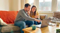 A man and a woman are seated on a brown leather couch, focused on a silver laptop on a wooden coffee table. The man, wearing a grey sweater, points at the laptop screen with a thoughtful expression, while the woman, dressed in a yellow sweater, holds a white mug and looks concerned. A bright orange knitted blanket is draped over the couch. A teal mug is on the coffee table, and a window showing residential houses in the background lets in bright sunlight.